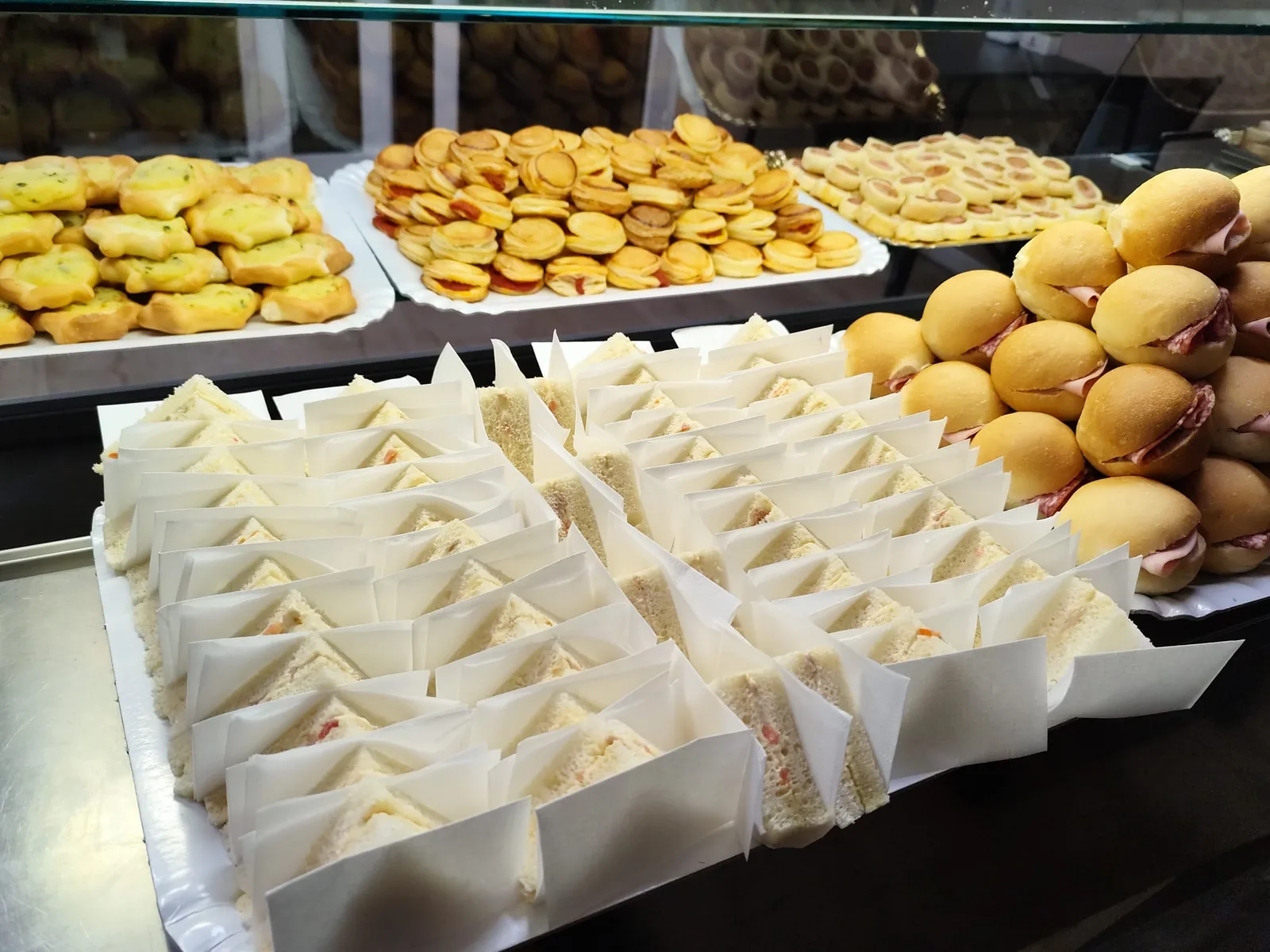 Banco allestito per un rinfresco: vassoi di tramezzini, panini mignon con affettati, sfogliatine e pardulas salate.
