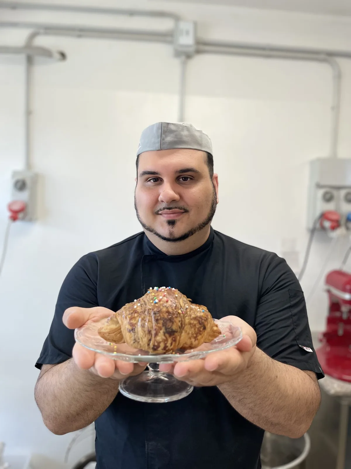 Le mani di Nicola Cara porgono un CroisSard glassato e decorato con confettini, su un'alzatina di vetro, in laboratorio.