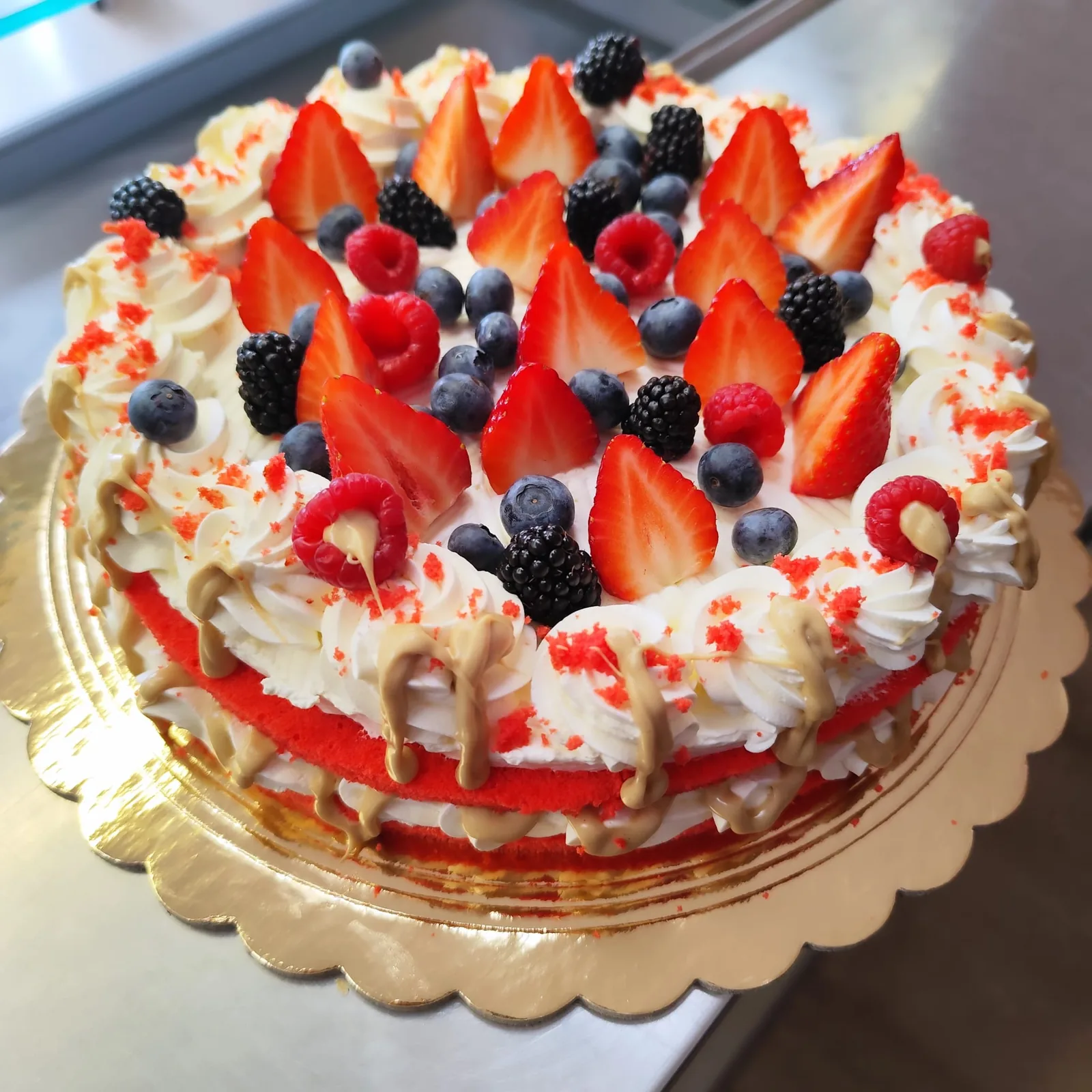 Torta tonda alla panna con fragole, lamponi, mirtilli e more, bordo di pan di Spagna rosso e colature di crema al caramello.