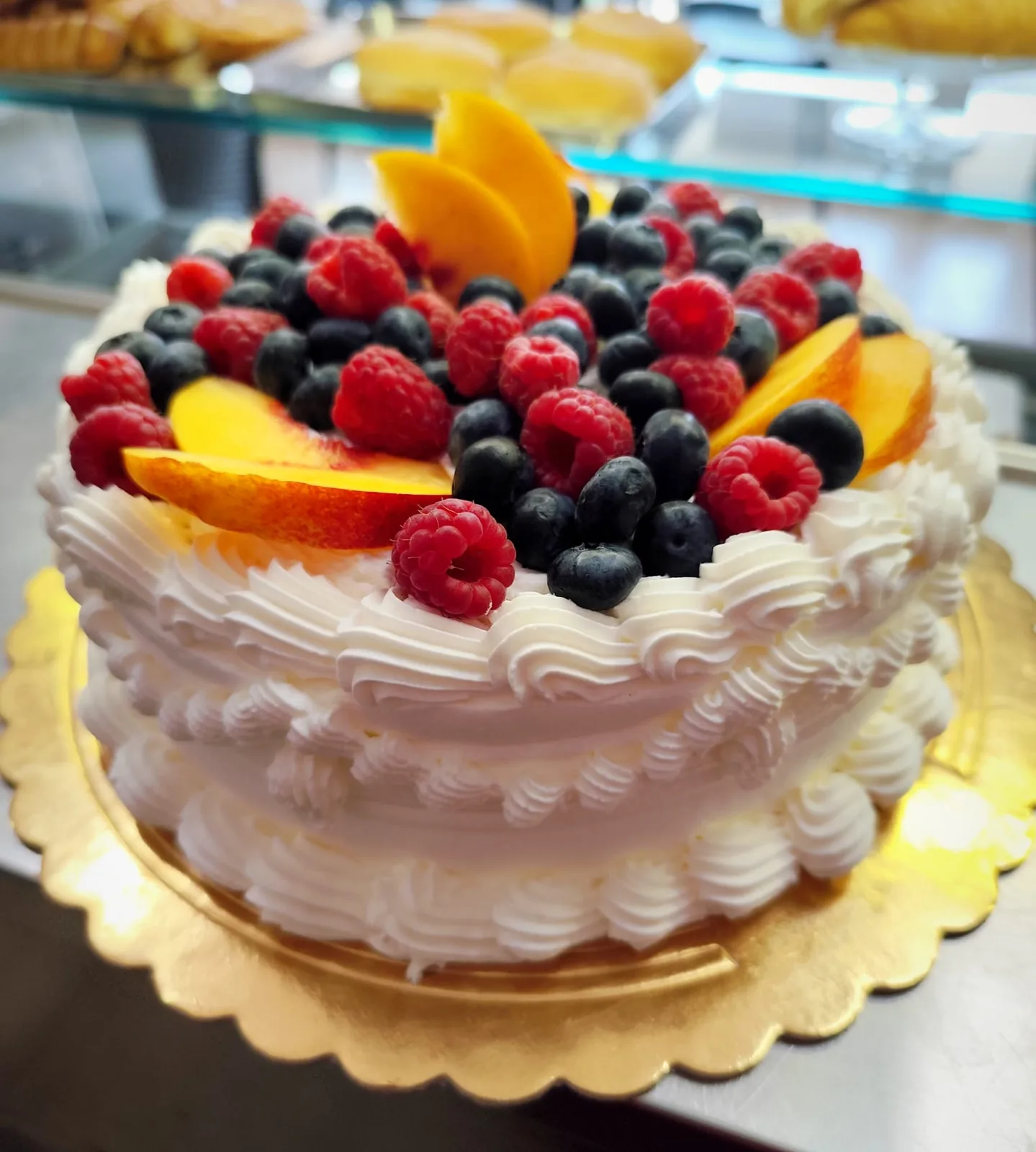Torta tonda ricoperta di panna montata a ciuffi, con frutti di bosco e spicchi di pesca al centro.